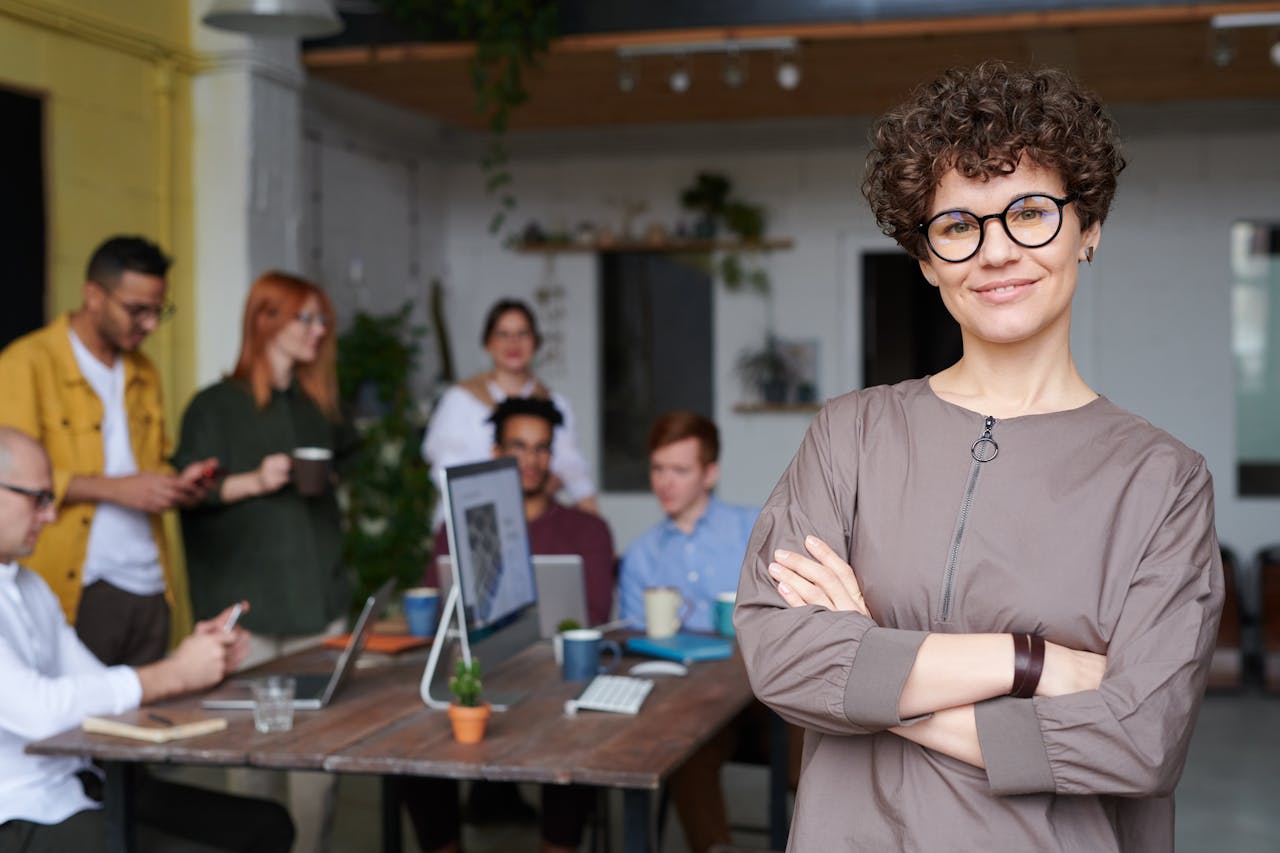 photo-of-woman-wearing-eyeglasses-3184405 Smiling businesswoman with curly hair stands confidently in a modern office space with colleagues.